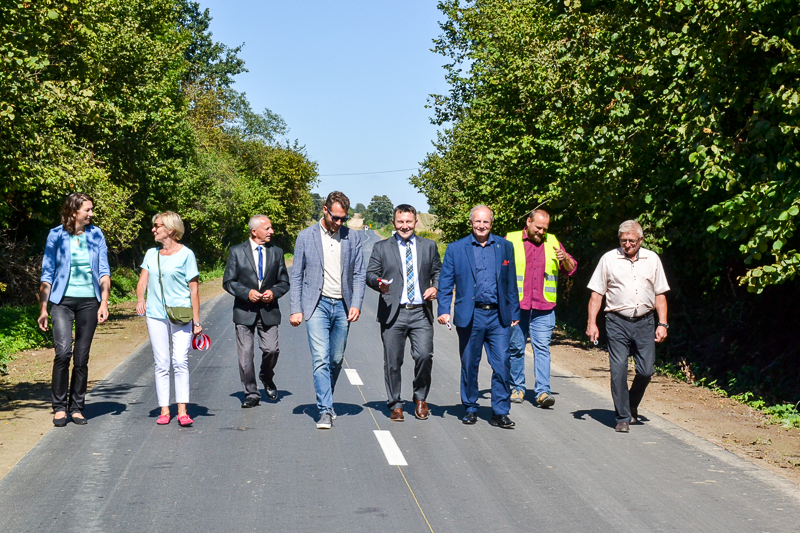 Grupa ośmiu osób idących po świeżo wyremontowanej, asfaltowej drodze otoczonej zielonymi drzewami pod jasnym, niebieskim niebem.