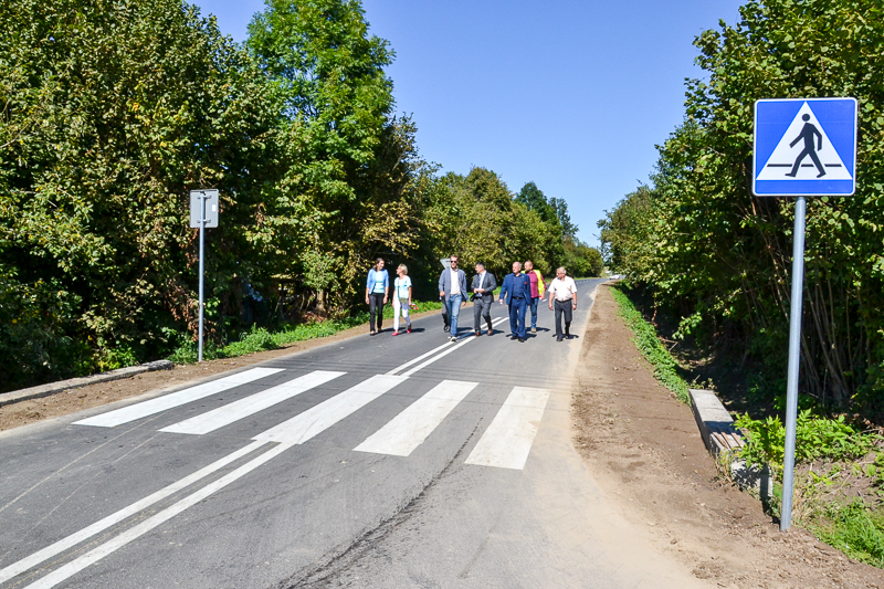 Grupa sześciu osób idących po przejściu dla pieszych na asfaltowej drodze otoczonej zielonymi drzewami pod błękitnym niebem.