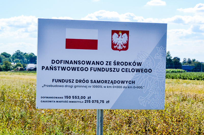Tablica informacyjna z flagą Polski, godłem oraz tekstem o dofinansowaniu przebudowy drogi gminnej, stojąca na tle zielonego pola i niebieskiego nieba z chmurami.