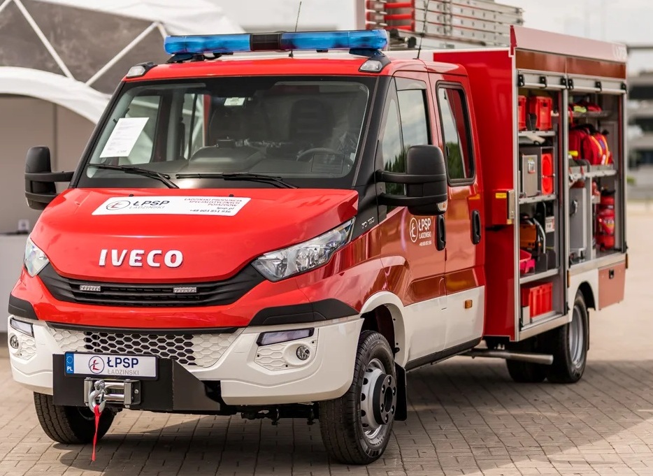 Czerwony i biały samochód strażacki Iveco z niebieskim światłem sygnalizacyjnym na dachu i otwartymi bocznymi schowkami pełnymi wyposażenia ratowniczego.