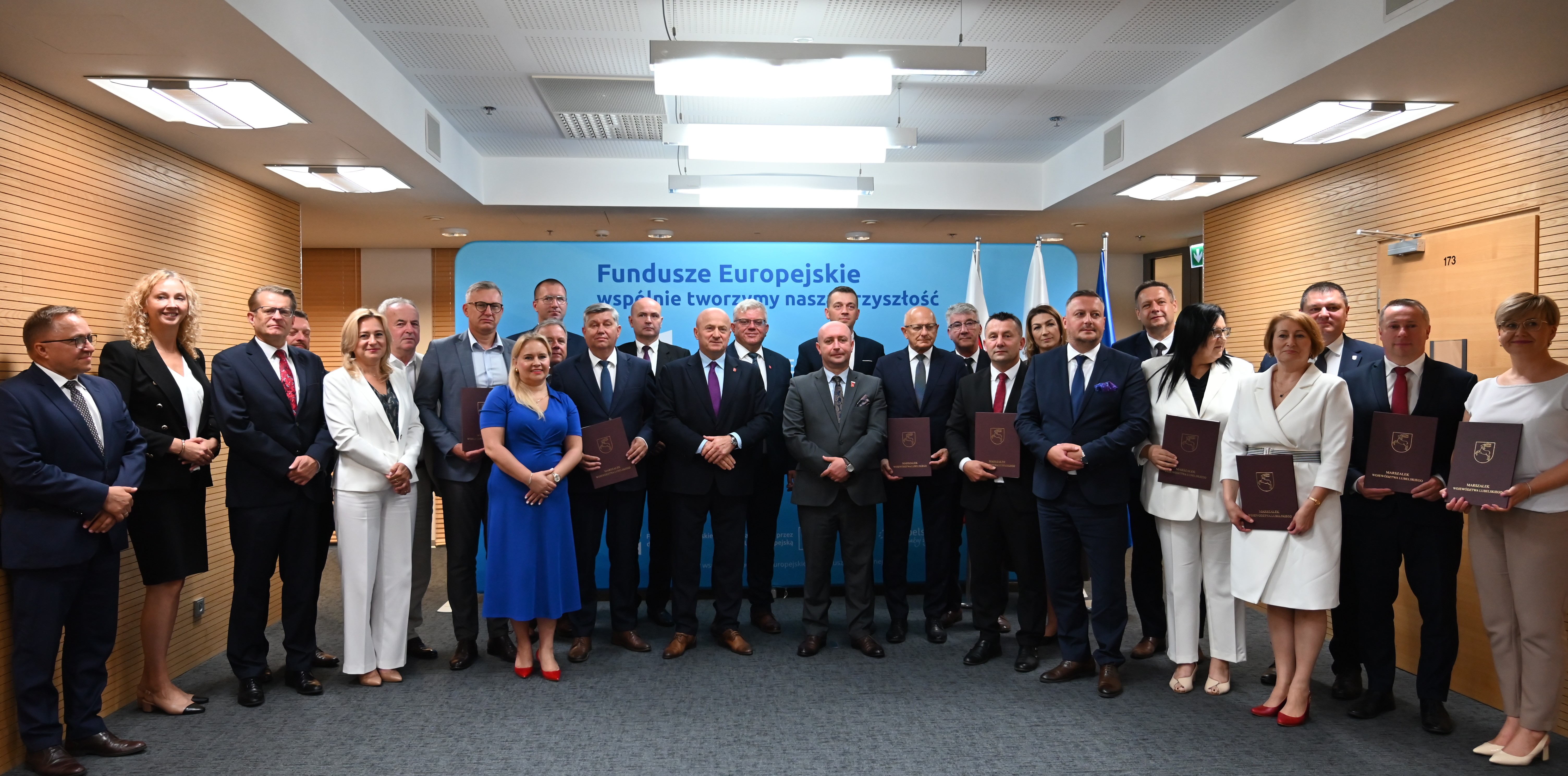 Grupa 25 osób w formalnych ubraniach stoi na tle niebieskiego banera z napisem Fundusze Europejskie, drewniane ściany, szare wykładziny, sufit z białymi panelami i lampami.