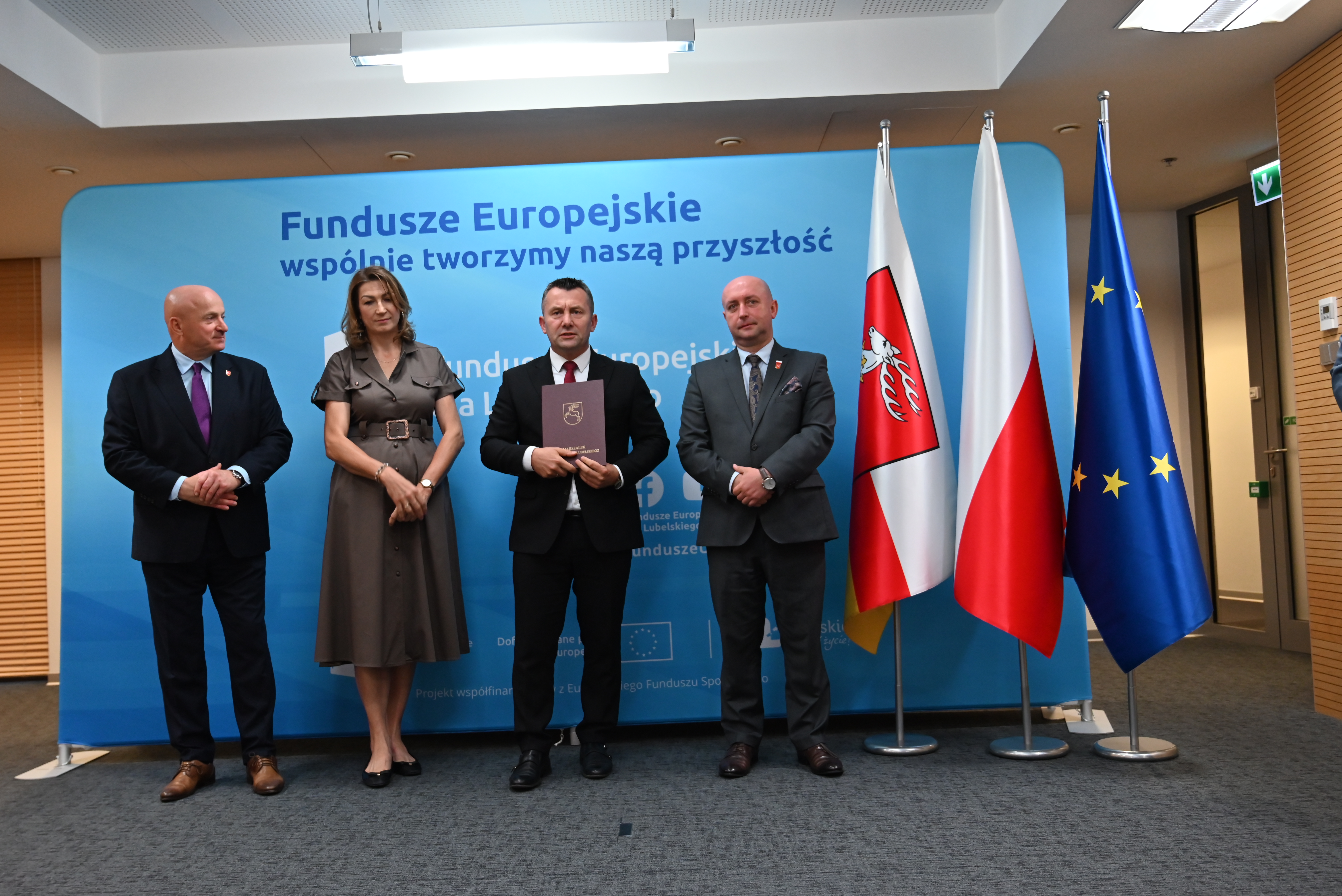 Cztery osoby w garniturach i sukience stoją na tle niebieskiego baneru z napisem Fundusze Europejskie, obok trzy flagi: z herbem, Polski i Unii Europejskiej.