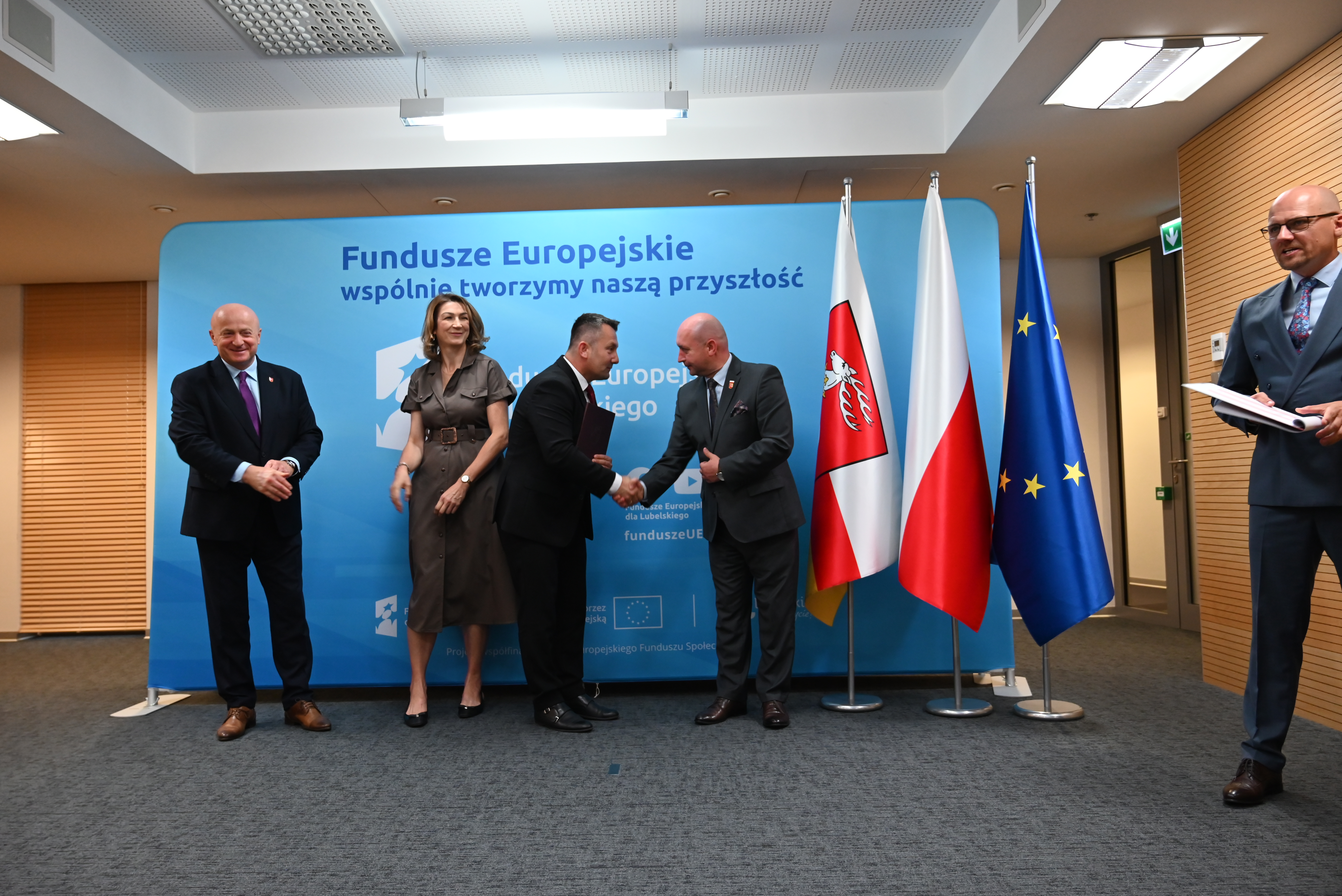 Pięć osób w garniturach i sukience stoi na szarym dywanie przed niebieskim ekranem z napisem, obok trzy flagi: z orłem, Polski i Unii Europejskiej.