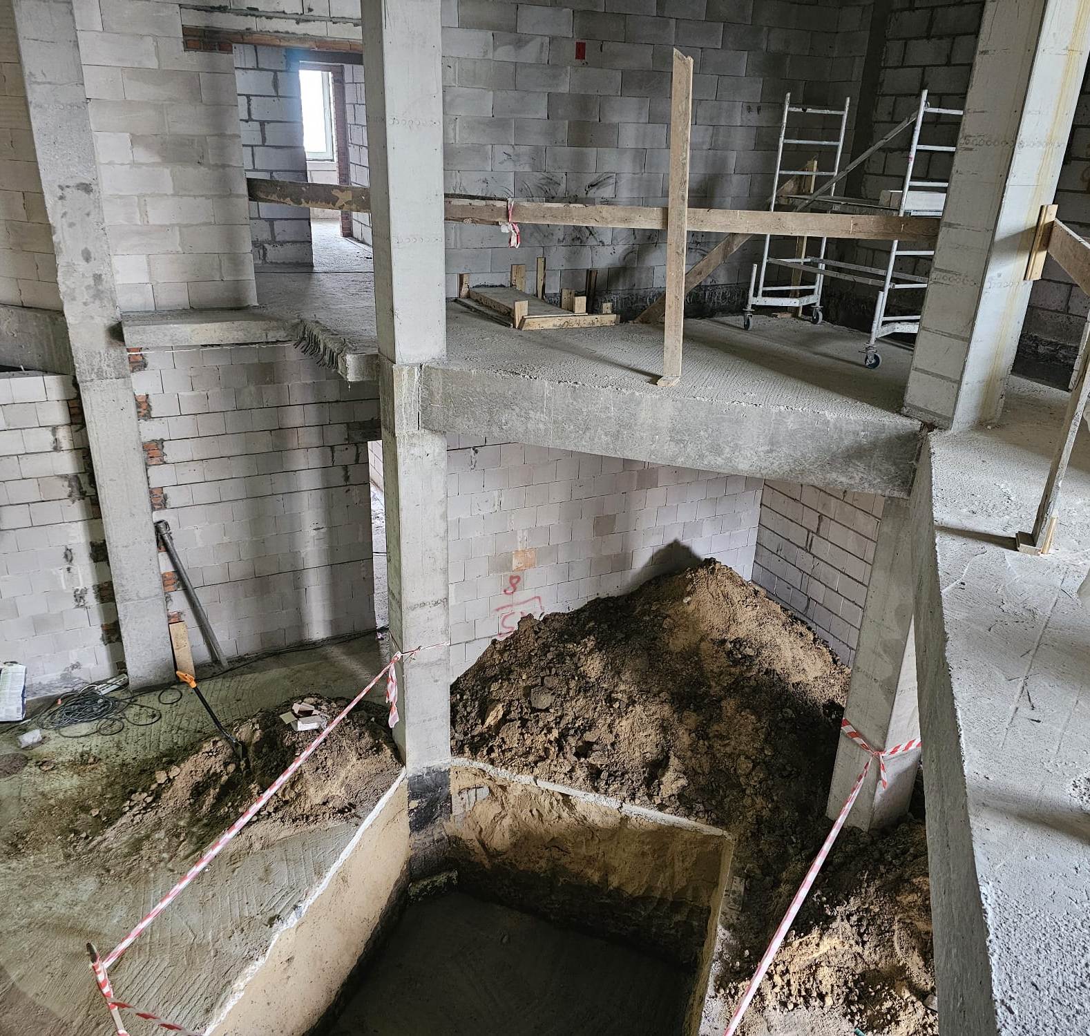 Wnętrze budynku w stanie surowym, betonowe ściany i podłogi, wykop ziemny zabezpieczony taśmą, stos ziemi, rusztowanie na kółkach, drewniane barierki ochronne.