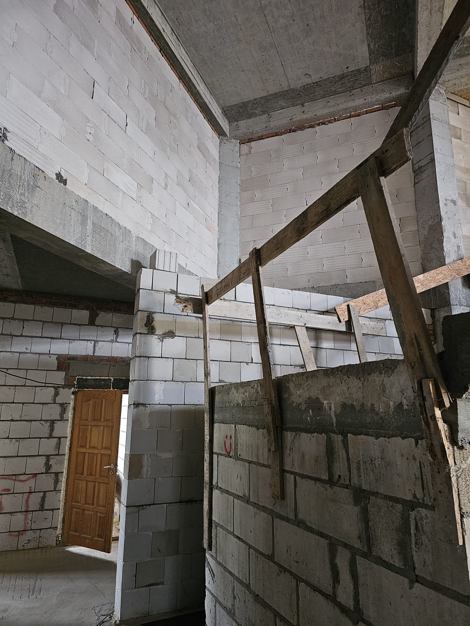 Wnętrze niedokończonego budynku z szarobiałymi ścianami z bloczków, drewnianym prowizorycznym balustradą i częściowo otwartymi drewnianymi drzwiami.