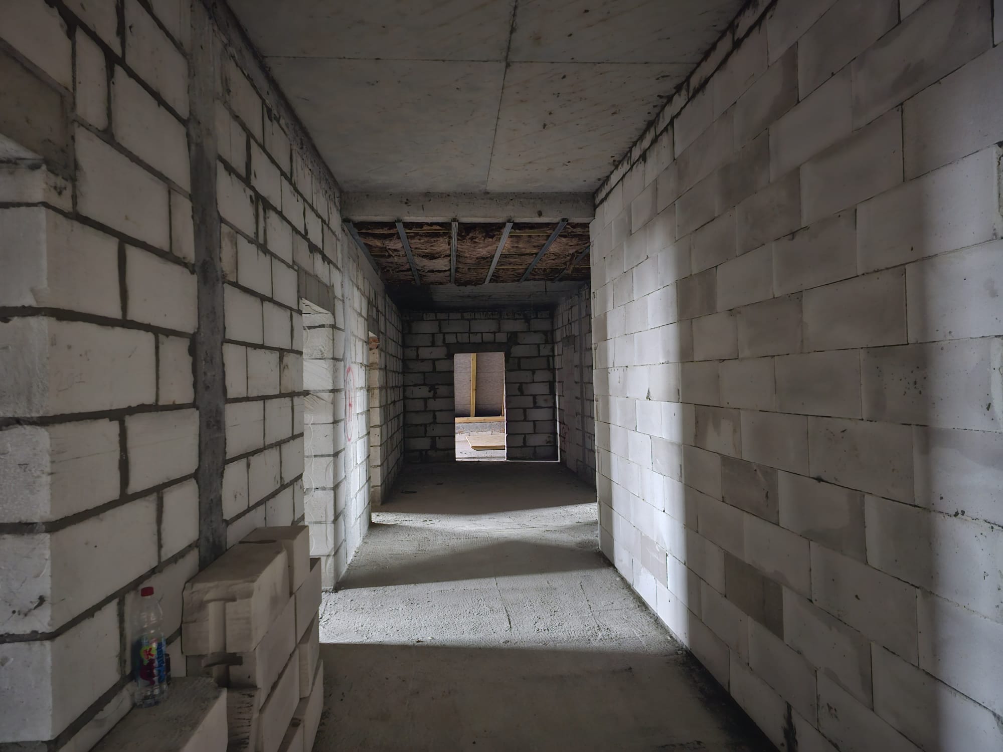 Wąski korytarz z białych bloczków, betonowa podłoga i sufit, stos bloczków po lewej stronie, butelka plastikowa na podłożu, światło padające z otworu na końcu korytarza.