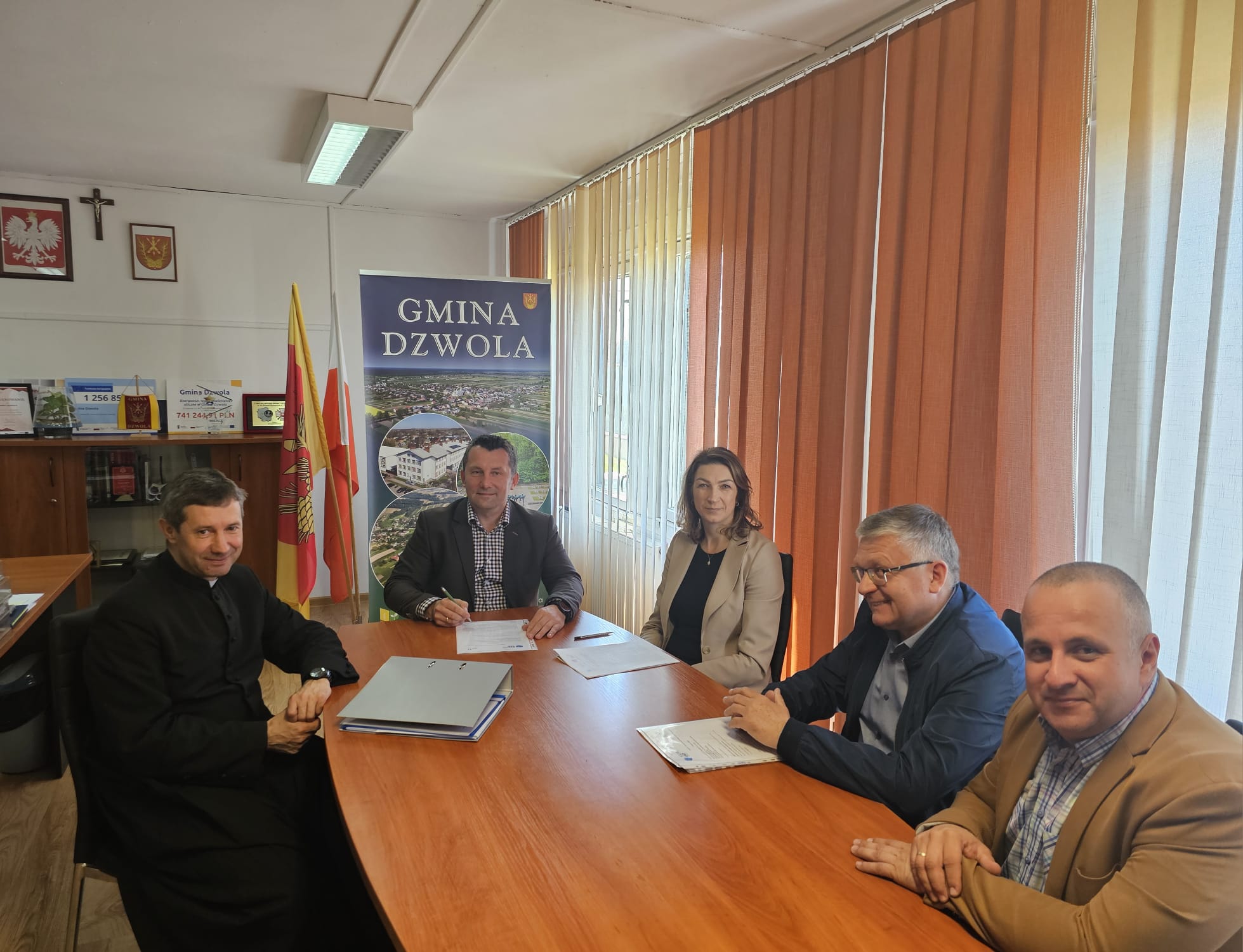 Pięć osób siedzi przy drewnianym stole w jasnym pomieszczeniu, za nimi flagi i banner z napisem "Gmina Dzwola", na ścianie krzyż i godło Polski.