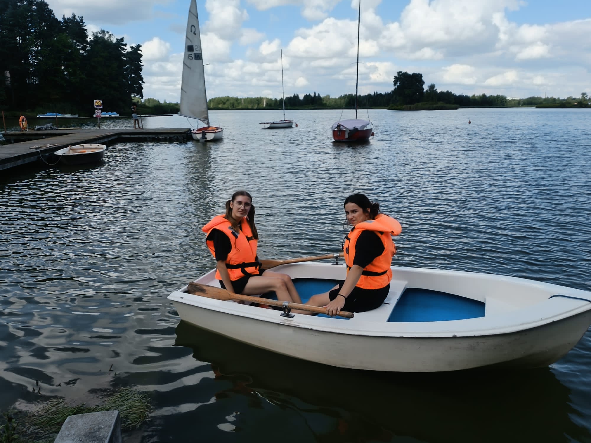 Dwie osoby w pomarańczowych kamizelkach ratunkowych siedzące w białej łodzi na jeziorze z żaglówkami i drewnianym pomostem w tle.