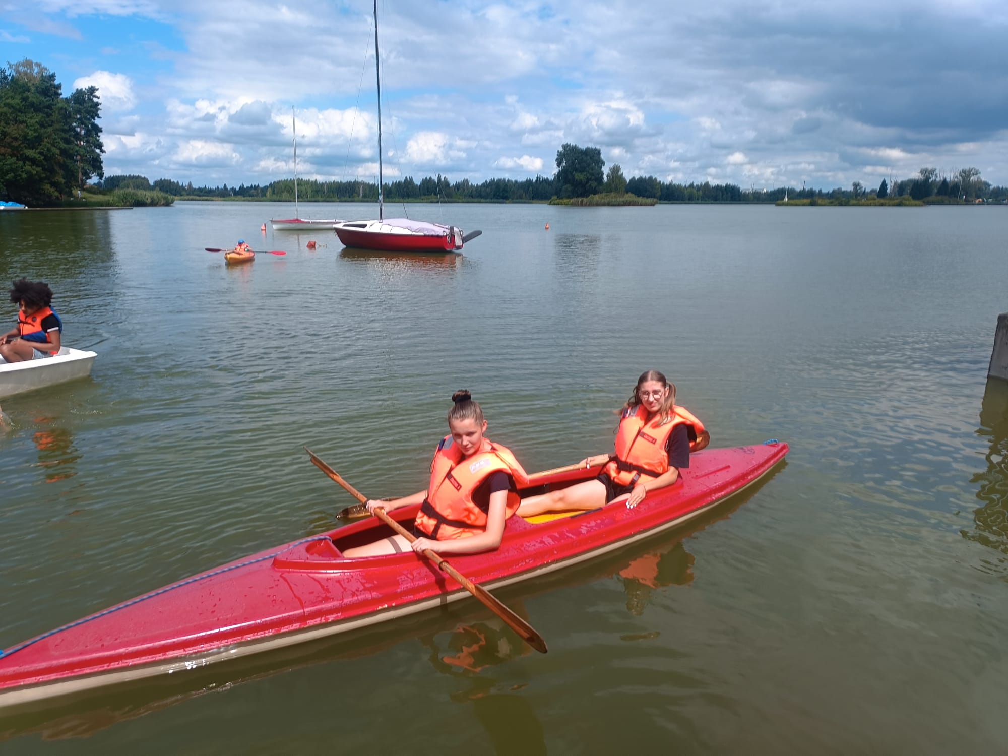 Dwie osoby w pomarańczowych kamizelkach siedzą w czerwonej kajakowej łodzi na jeziorze, w tle żaglówki i drzewa pod pochmurnym niebem.