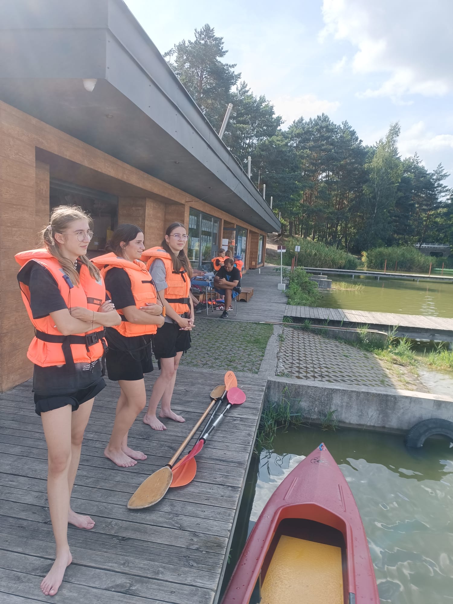 Trzy kobiety w pomarańczowych kamizelkach ratunkowych stoją boso na drewnianym pomoście obok czerwonego kajaka i trzech wioseł, w tle budynek i jezioro z drzewami.