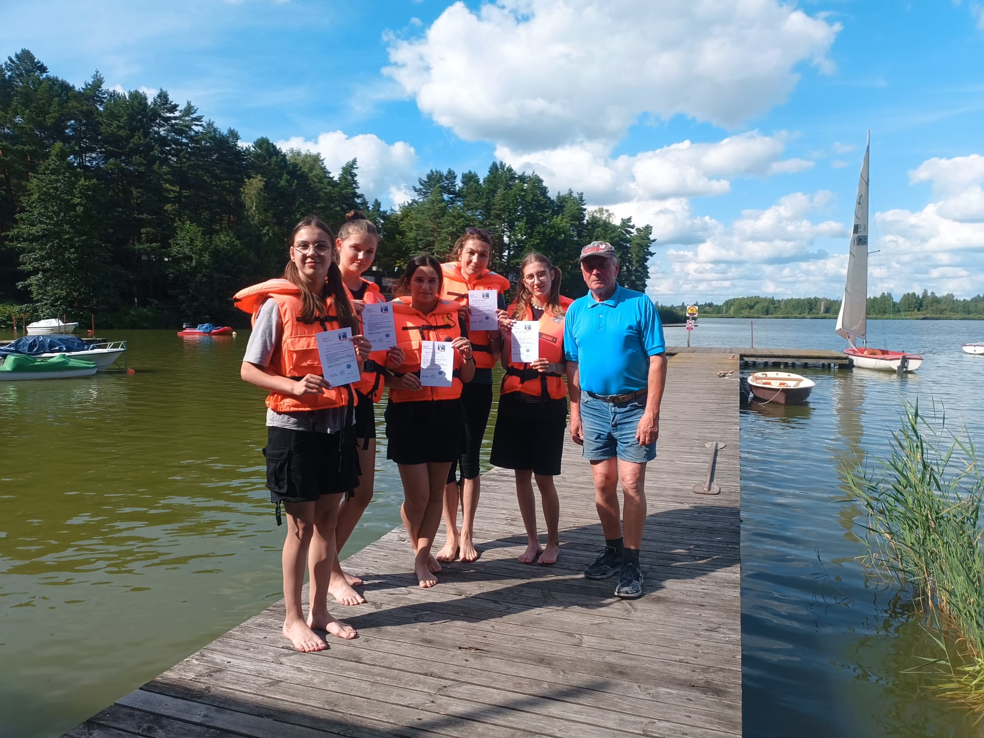 Grupa pięciu kobiet w pomarańczowych kamizelkach ratunkowych i mężczyzna w niebieskiej koszulce i szortach stoi na drewnianym pomoście nad jeziorem z żaglówkami i zielonym lasem w tle.