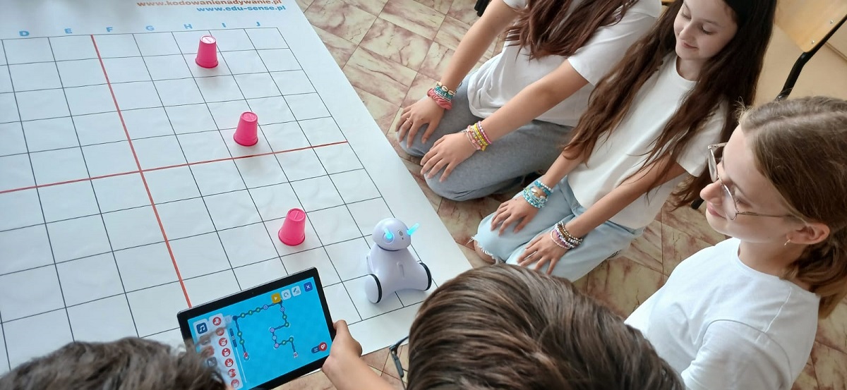 Cztery dzieci w białych koszulkach wokół maty z siatką, na której stoją trzy różowe kubki i mały biały robot, jedno dziecko trzyma tablet z aplikacją.