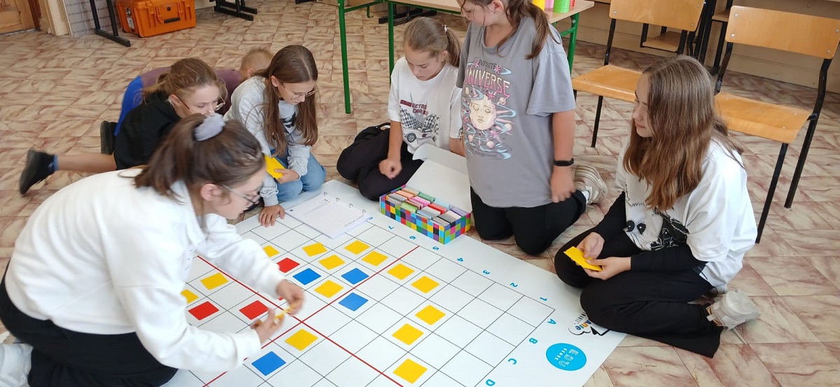 Siedem dzieci na podłodze wokół dużej planszy z kratkami, na której są ułożone żółte, czerwone i niebieskie kwadraty. W tle krzesła i stoły.