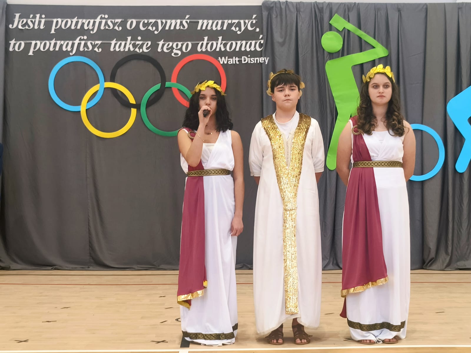 Trzy osoby w strojach greckich z wieńcami laurowymi, dwie kobiety w białych i bordowych togach, chłopak w białej todze z złotą lamówką, na tle szarych zasłon i symboli olimpijskich.