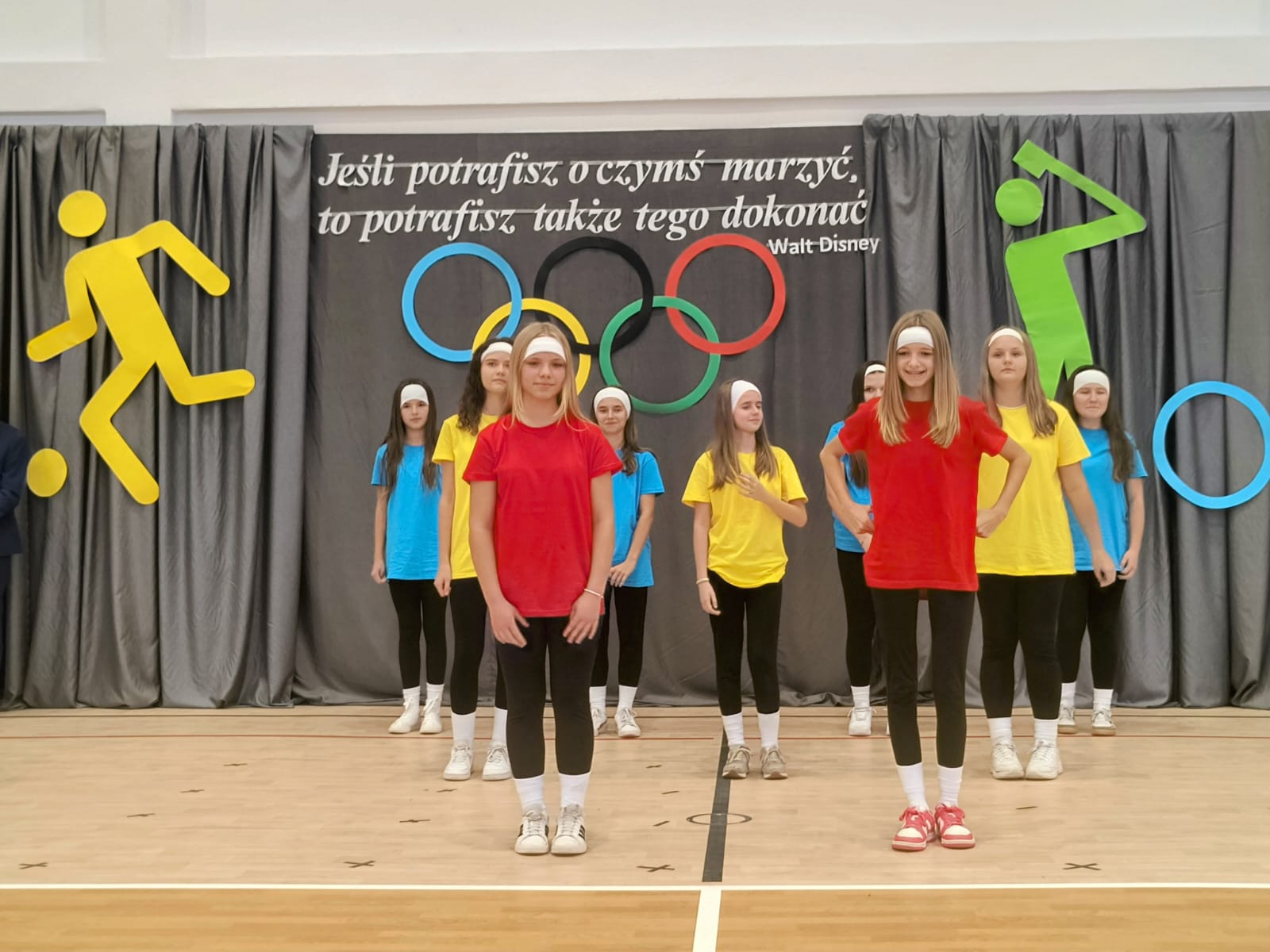 Dziewczęta w kolorowych koszulkach (czerwone, żółte, niebieskie), czarnych legginsach i białych opaskach na głowach stoją na drewnianej sali gimnastycznej na tle szarych zasłon z kolorowymi symbolami i napisem.