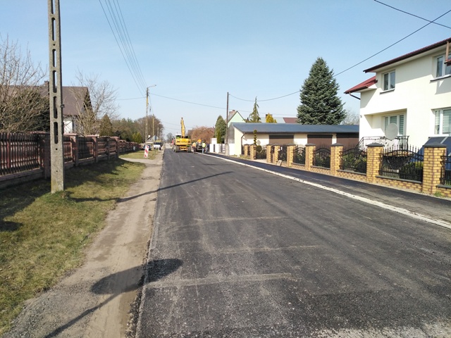 Ulica z nowo położonym asfaltem, z lewej strony trawnik i drewniany płot, z prawej dom z białą elewacją i żółtym ogrodzeniem z cegieł, w tle maszyna budowlana.