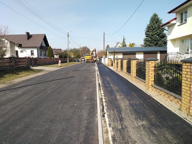 Ulica z nowo położonym ciemnym asfaltem, po prawej stronie żółto-brązowy mur z metalowym ogrodzeniem, po lewej domy jednorodzinne i zielone drzewa, jasne niebo.