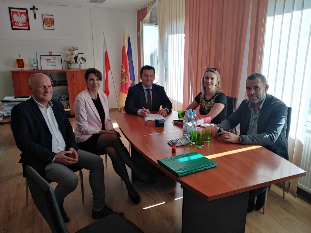 Pięć osób siedzi przy drewnianym stole w jasnym biurze z dużymi oknami i pionowymi żaluzjami, na ścianie flagi i godła, na stole butelki wody, szklanki i segregatory.