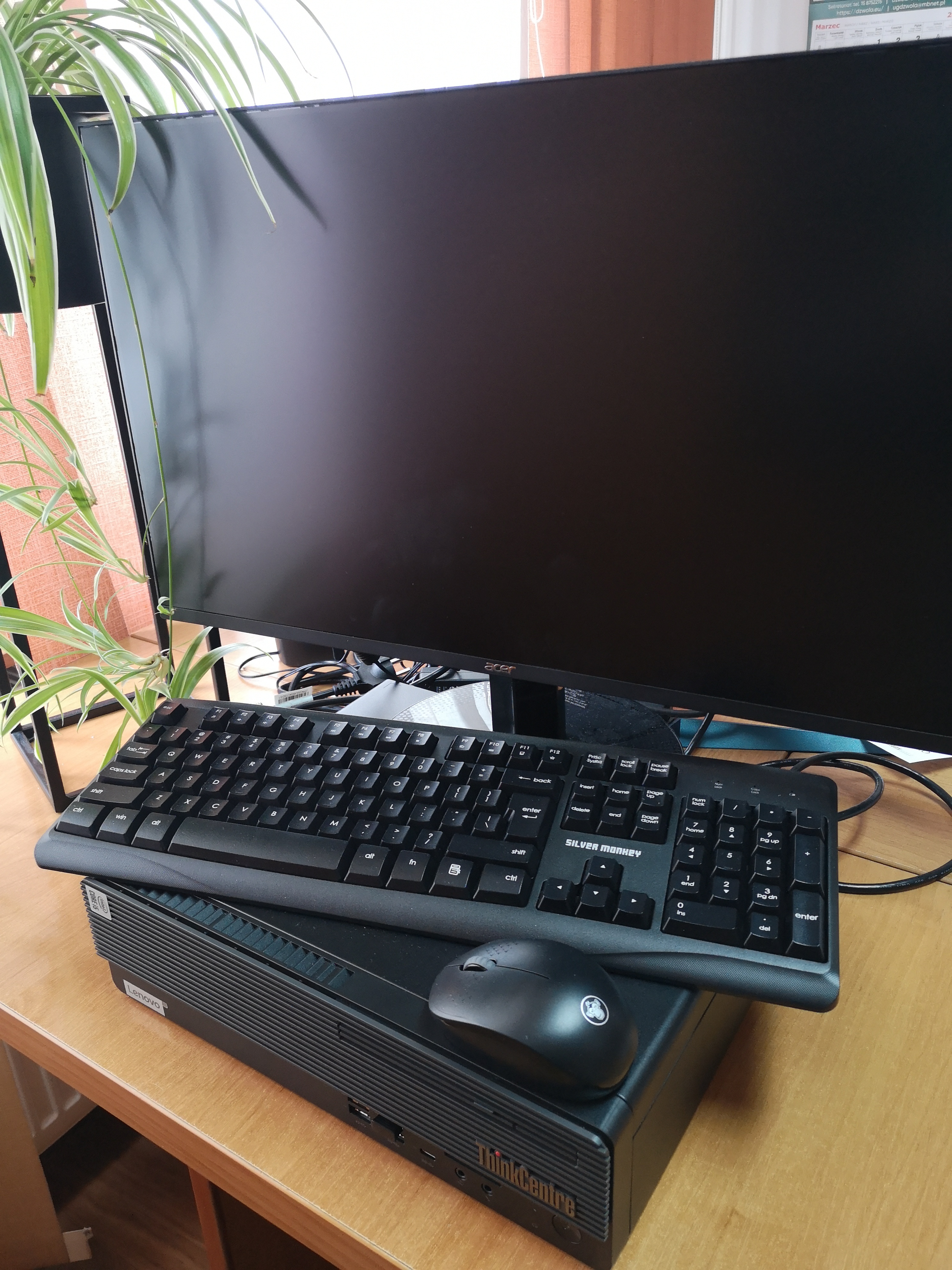 Monitor Acer z czarnym wyłączonym ekranem, na nim czarna klawiatura Silver Monkey i myszka, pod nimi czarna obudowa Lenovo ThinkCentre na drewnianym biurku, obok zielone liście rośliny.