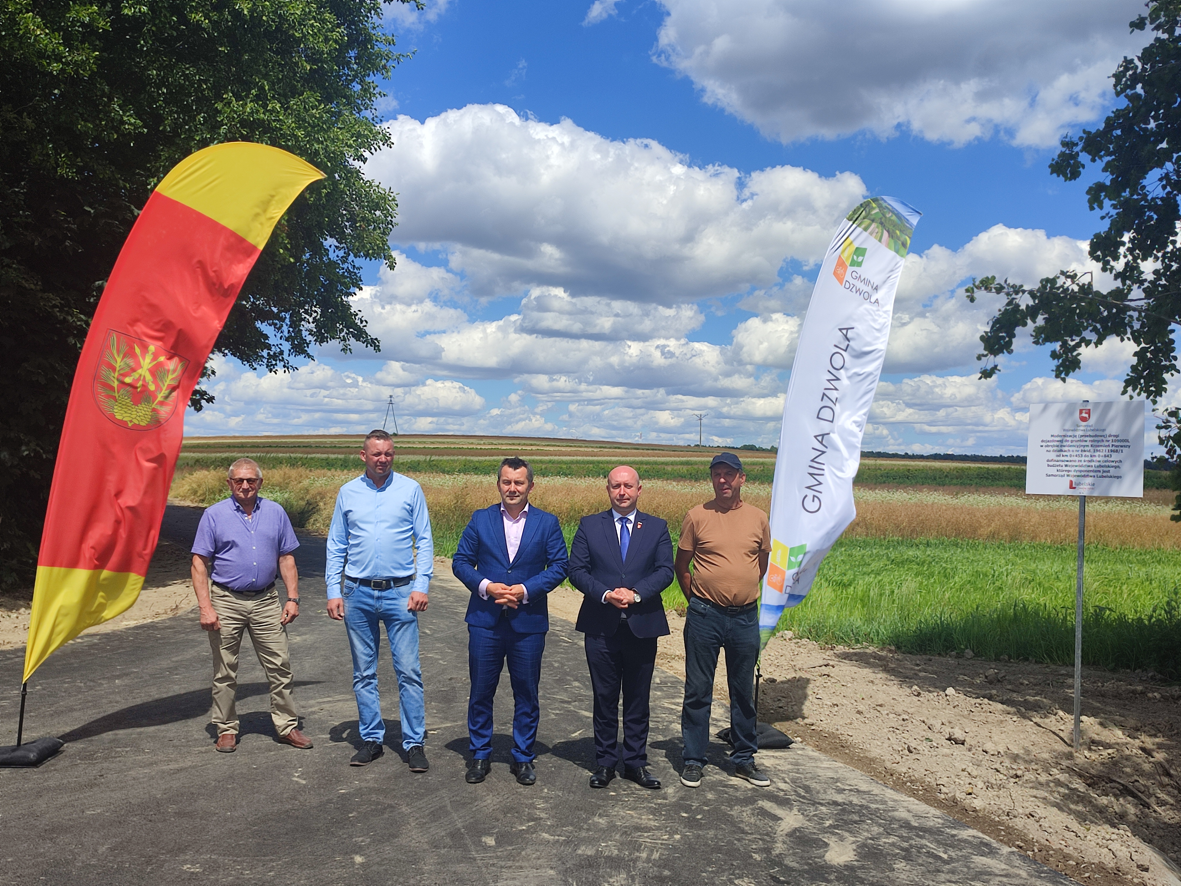 Pięciu mężczyzn stoi na świeżo wyasfaltowanej drodze, po bokach flagi: czerwona z żółtym i herbem oraz biała z napisem "Gmina Dzwola", w tle pola i niebo z chmurami.