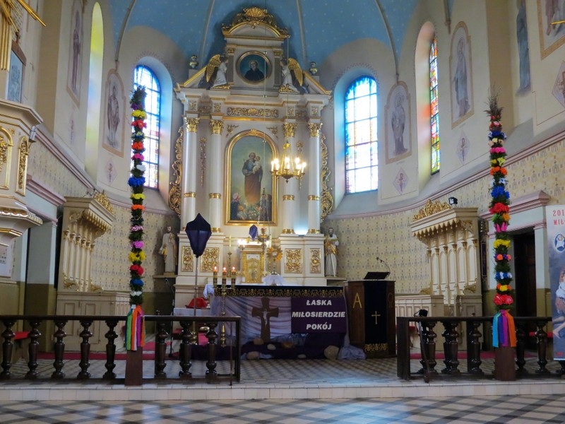 Wnętrze kościoła z ołtarzem ozdobionym złotymi detalami, niebieskim sklepieniem i dwoma wysokimi kolorowymi palmami po bokach.