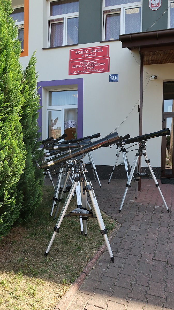 Sześć czarnych teleskopów na srebrnych statywach ustawionych na trawie i chodniku przed budynkiem z białą elewacją i czerwonymi tablicami.