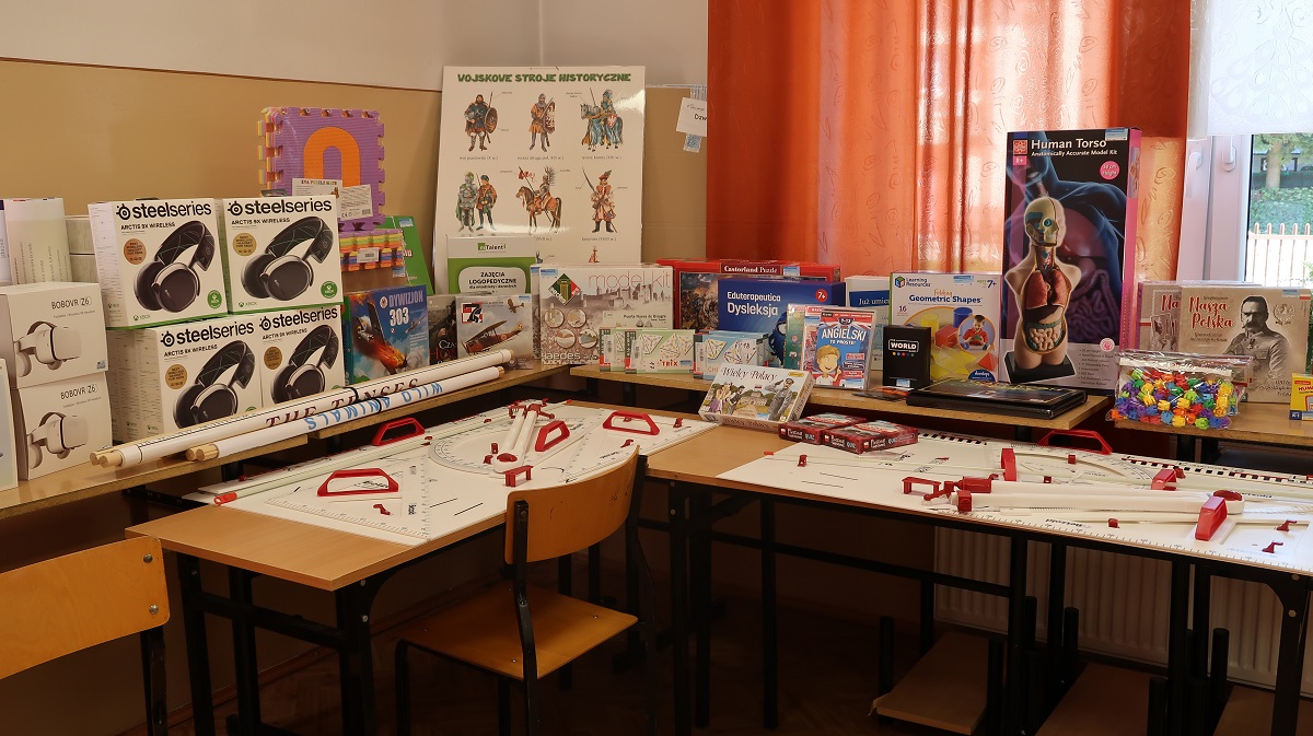 Zestaw edukacyjnych gier, modeli i słuchawek na stołach w klasie z pomarańczowymi zasłonami i drewnianymi krzesłami.