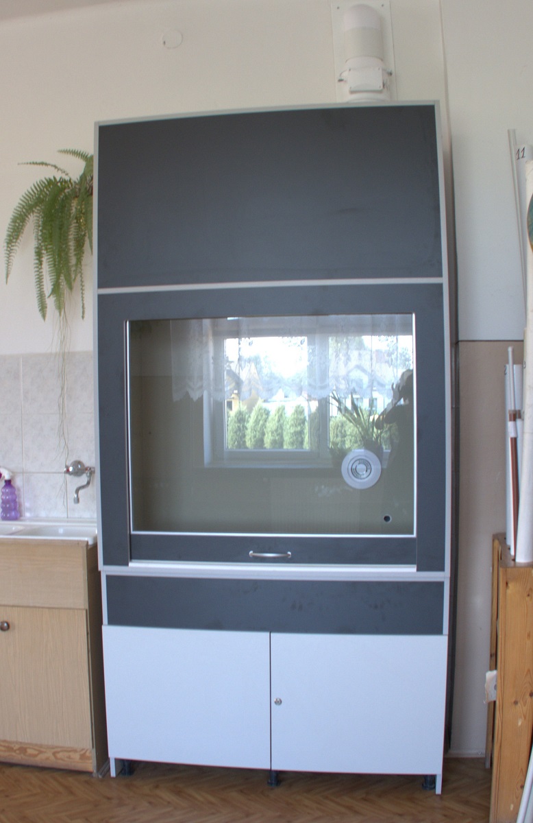 Szafka laboratoryjna w kolorze czarnym i białym z dużym przezroczystym ekranem, na którym widać odbicie okna i roślin, obok zlew z kranem i wisząca paprotka.