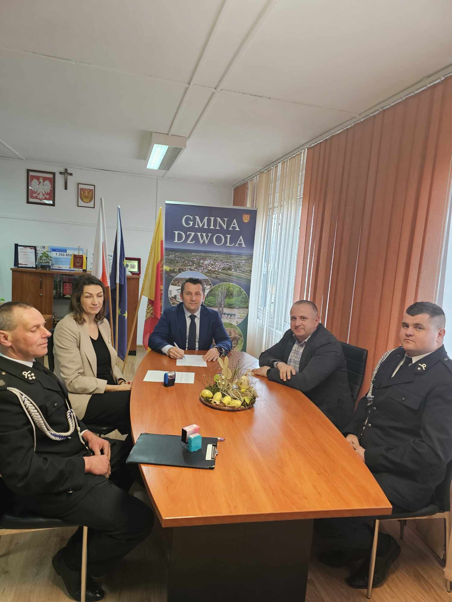 Pięć osób siedzi przy drewnianym stole w biurze z flagami i banerem "Gmina Dzwola", na stole dekoracja wielkanocna z jajkami i króliczkiem.