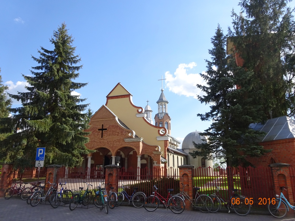 Ceglana zabudowa z elementami beżu, krzyże na budynkach, rowery zaparkowane przy czerwonym ogrodzeniu, wysokie drzewa iglaste, niebieskie niebo z chmurami.