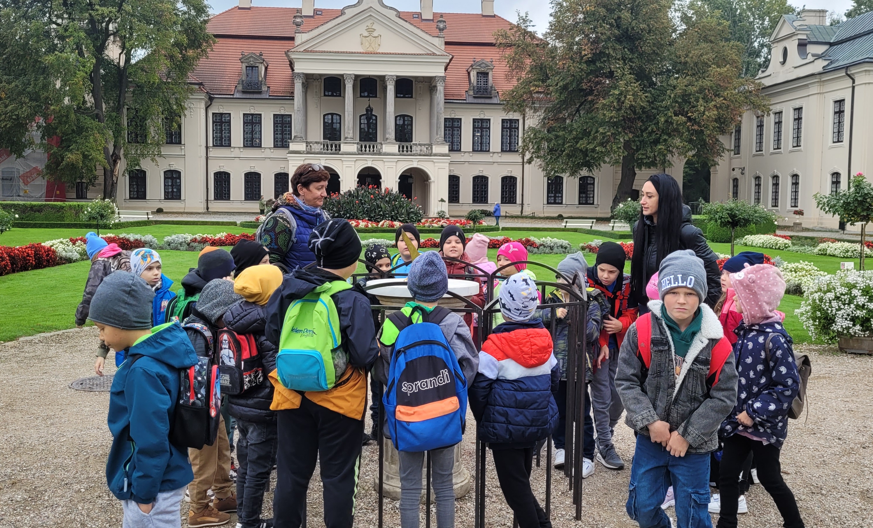 Grupa dzieci w kolorowych kurtkach i czapkach stoi przy metalowej konstrukcji na dziedzińcu z zieloną trawą, kwiatami i historycznym budynkiem w tle.
