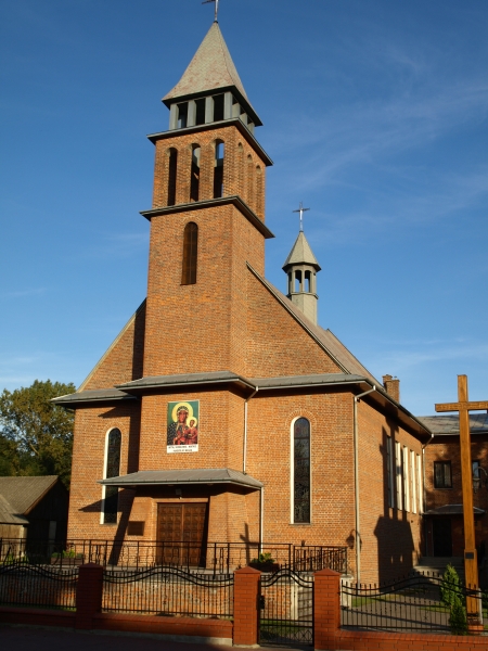 Ceglana świątynia z dwoma wieżami i krzyżami, wmurowany obraz Matki Boskiej z Dzieciątkiem, jasno niebieskie niebo, metalowy płot z ceglaną podmurówką.