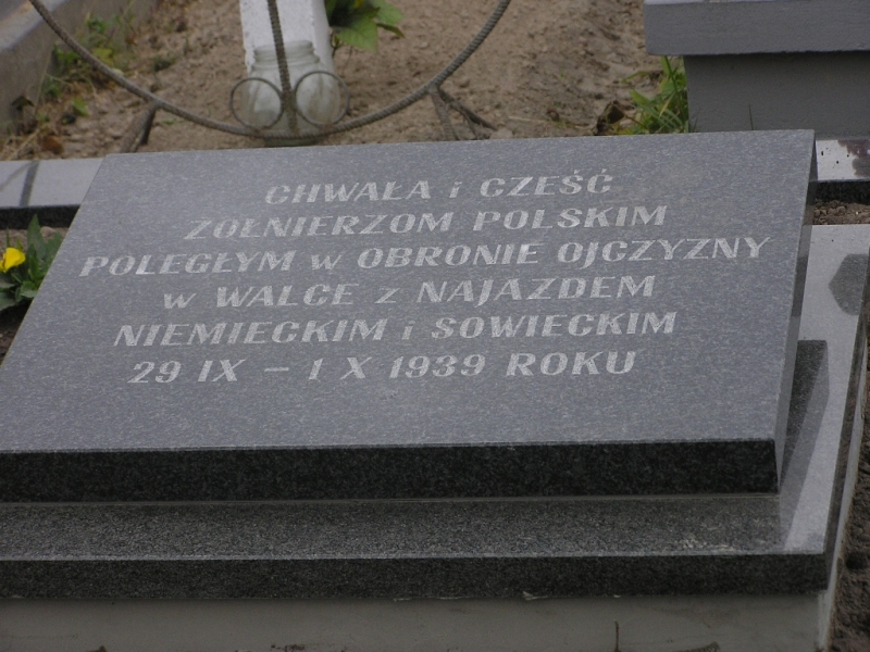 Czarna płyta nagrobna z białym napisem w języku polskim na tle ziemi i części płotu.