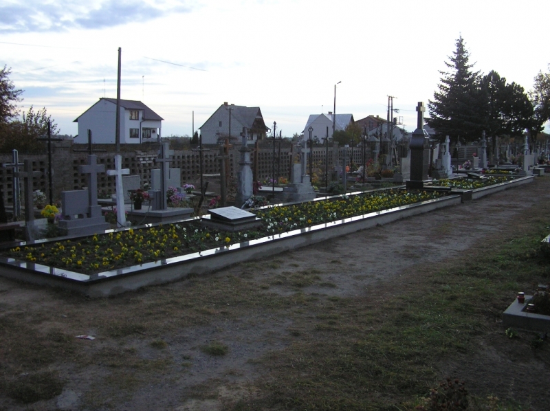 Nagrobki i groby z kwiatami na cmentarzu, w tle niskie domy i drzewa pod zachmurzonym niebem.