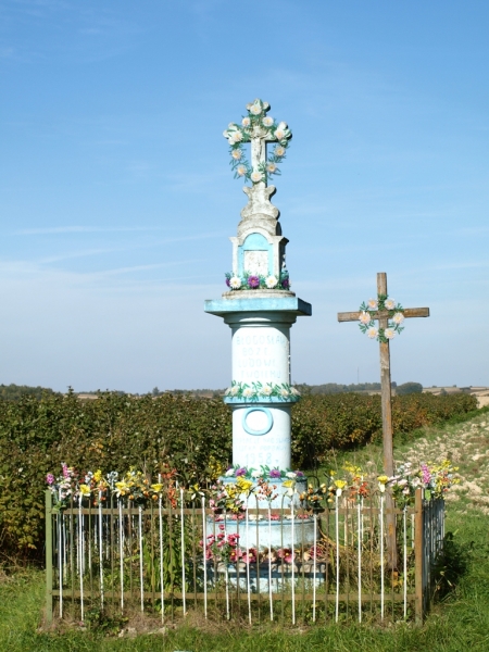 Kamienny biały krzyż i drewniany krzyż z ozdobami kwiatowymi na tle pola i błękitnego nieba, otoczone metalowym ogrodzeniem z kolorowymi sztucznymi kwiatami.