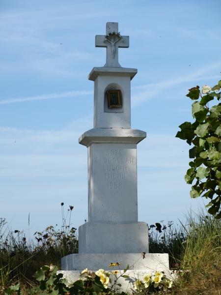 Biały kamienny krzyż z wizerunkiem ukrzyżowanego Chrystusa, stojący na kamiennym cokole, otoczony zielenią i kwiatami, na tle niebieskiego nieba.