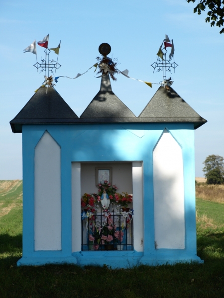 Niebieska kapliczka z trzema szczytami i ozdobnymi metalowymi krzyżami na dachu, w otwartej wnęce figura Matki Boskiej otoczona sztucznymi kwiatami, zielone pole w tle.