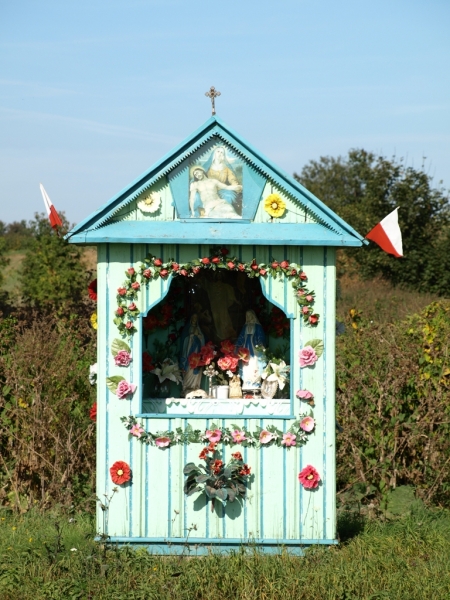 Niebieska kapliczka z dekoracją z różowych i czerwonych kwiatów, obrazem religijnym na szczycie i dwoma białymi flagami z czerwonym paskiem na tle zielonej roślinności.