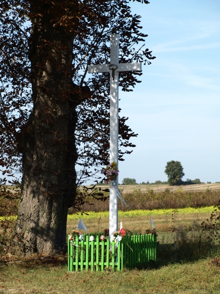 Biały krzyż z kwiatami i wstążkami przy dużym drzewie, otoczony niskim zielonym ogrodzeniem, na tle pola i błękitnego nieba.