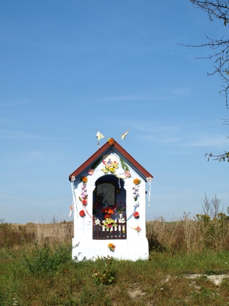Biała kapliczka z czerwoną dachówką, ozdobiona kolorowymi kwiatami i girlandami, stoi na tle niebieskiego nieba i suchej trawy.