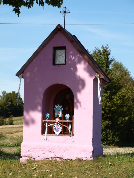 Różowa kapliczka z daszkiem, metalowy krzyż na szczycie, ozdoby z białych wstążek, na tle zielonych drzew i błękitnego nieba.