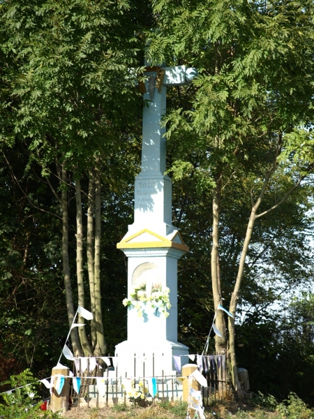 Biały kamienny krzyż z żółtym daszkiem na postumencie, otoczony niskim ogrodzeniem i girlandami, na tle zielonych drzew.