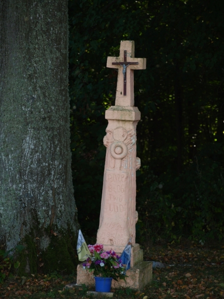 Różowy kamienny krzyż z metalowym Jezusem na tle ciemnozielonych drzew, u podstawy doniczka z kwiatami i dwie małe flagi.