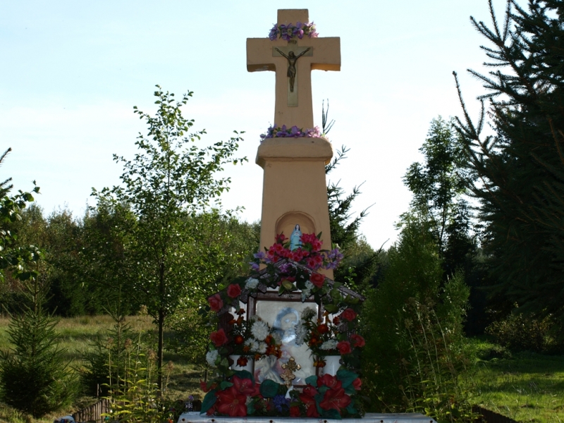 Beżowy kamienny krzyż z metalową figurą Jezusa, ozdobiony fioletowymi i czerwonymi kwiatami, na tle zielonych drzew i błękitnego nieba.