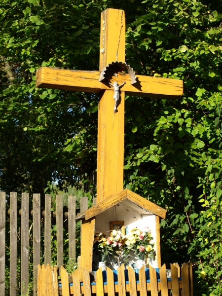 Żółty drewniany krzyż z metalową figurą Jezusa, pod nim niewielka kapliczka z kwiatami, otoczona niskim drewnianym płotkiem, w tle zielone liście drzew.