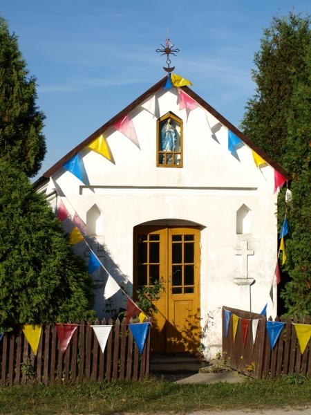 Biała kaplica z brązowymi drzwiami, pomiędzy dwoma zielonymi drzewami, udekorowana kolorowymi trójkątnymi flagami na tle błękitnego nieba.