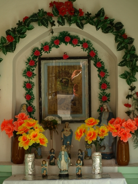 Kapliczka z figurami Matki Boskiej i dzieci, otoczona sztucznymi kwiatami w czerwono-żółtych barwach oraz girlandą z liści i róż. Wewnątrz obraz w ramie.