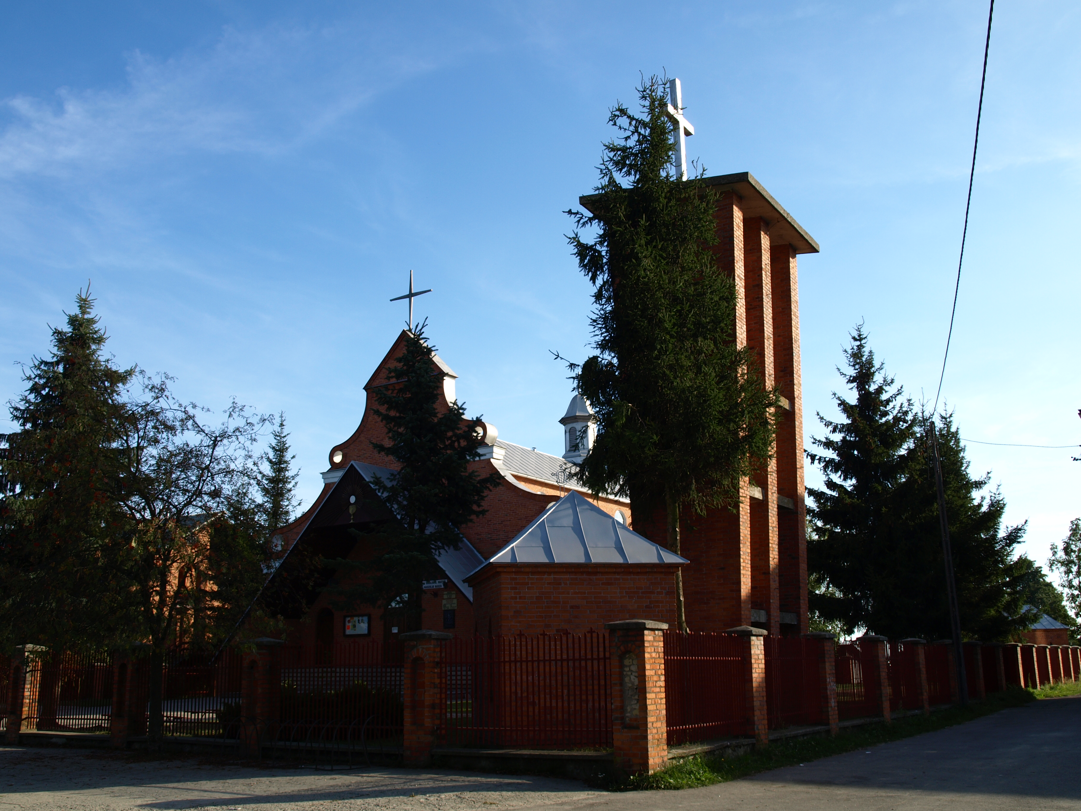 Ceglany kościół z wieżą i metalowym dachem, otoczony zielonymi drzewami i płotem z cegły i metalu na tle niebieskiego nieba.