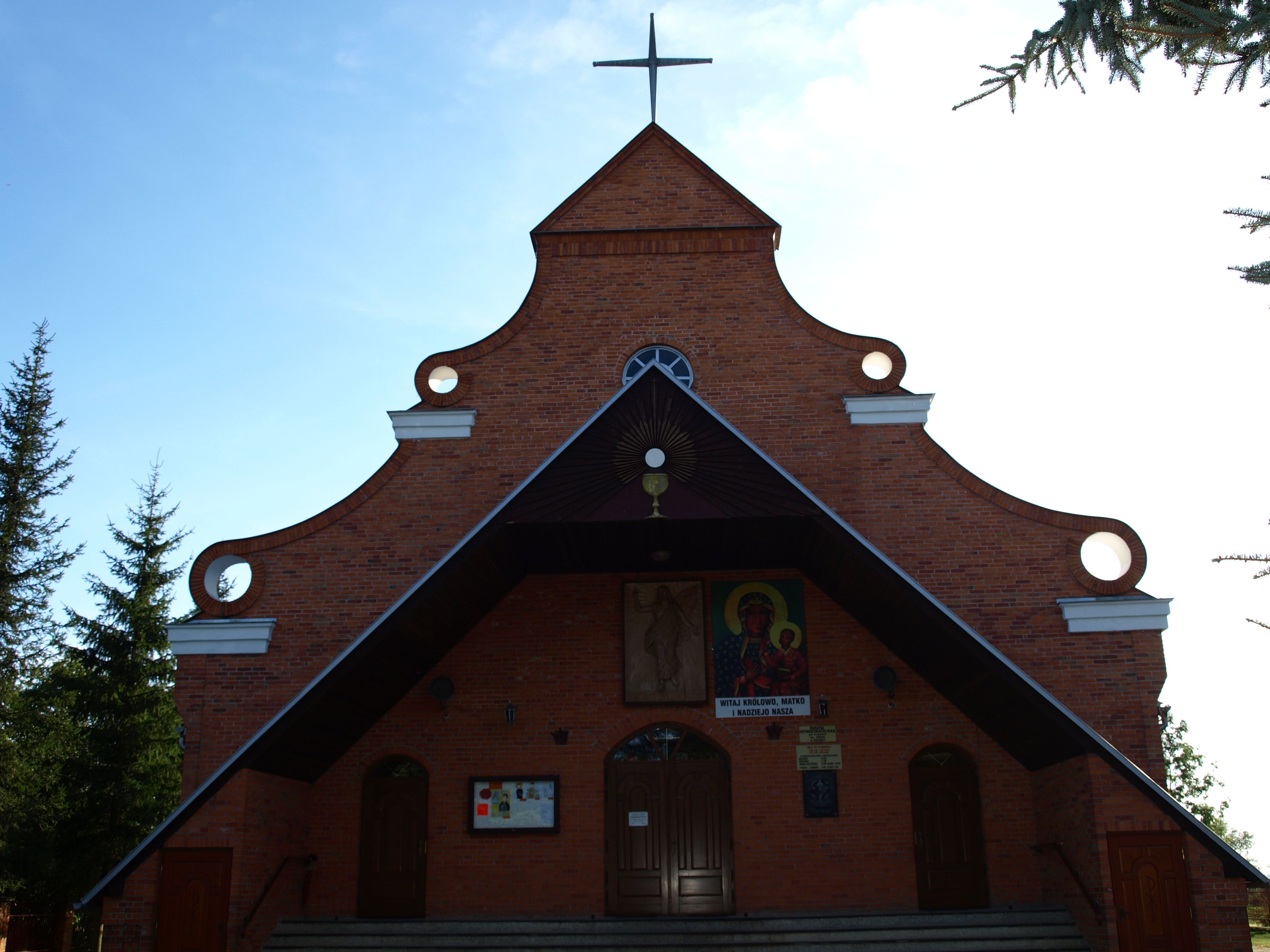 Ceglany front kościoła z dwoma wejściami, dużym daszkiem w kształcie trójkąta, krzyżem na szczycie i obrazami nad drzwiami, otoczony drzewami na tle jasnego nieba.