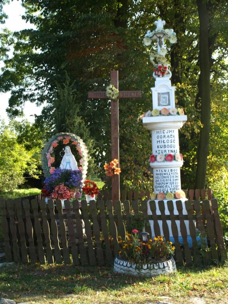 Ołtarzyk polowy z białą figurą Matki Boskiej otoczoną wieńcem kwiatów, drewniany ciemny krzyż z napisem, biały postument z krzyżem i tabliczkami z tekstem, ogrodzony drewnianym płotem.
